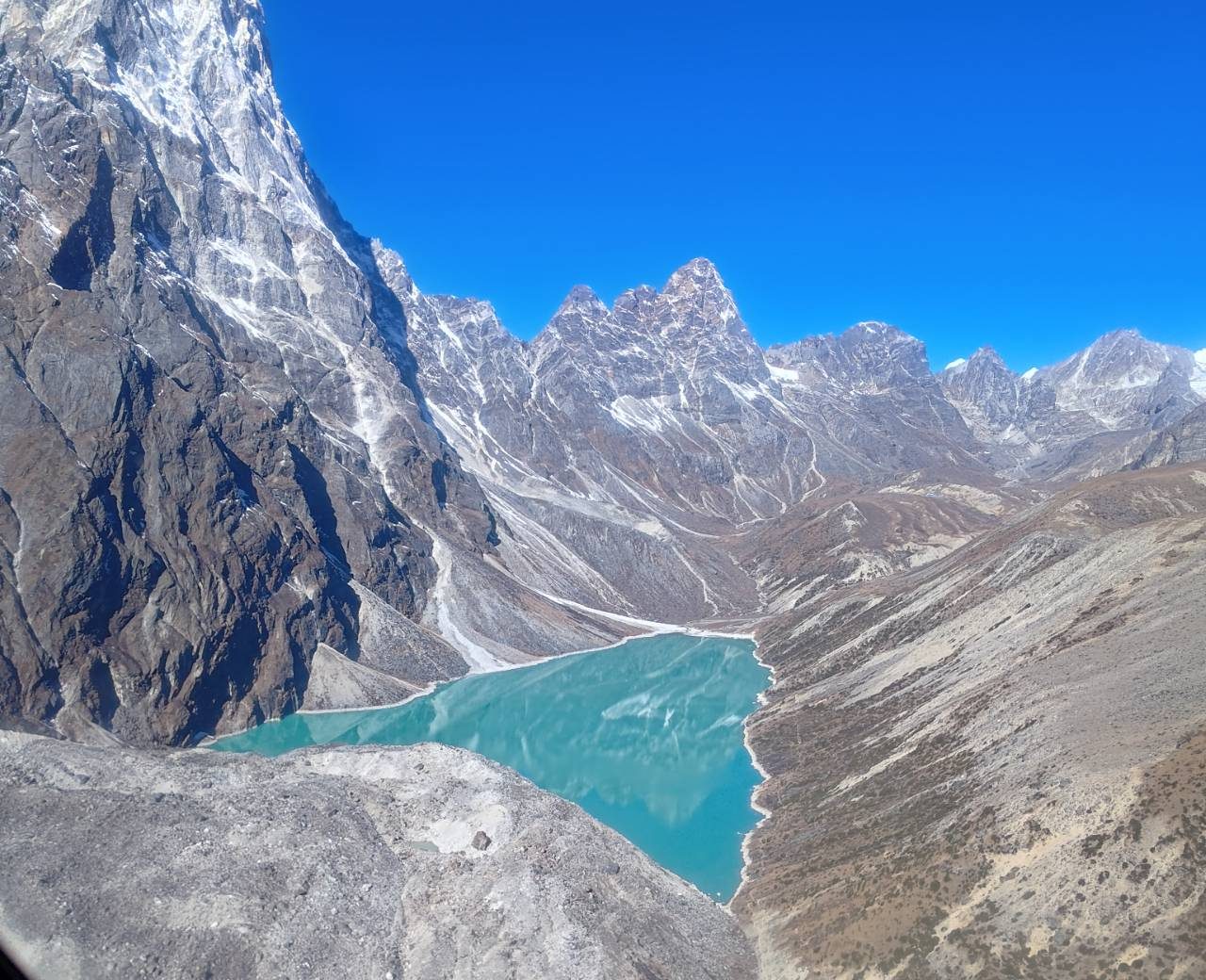 A turquoise glacial lake surrounded by steep rocky mountains and snow peaks, viewed from above during the Everest Base Camp Helicopter Tour in Nepal.