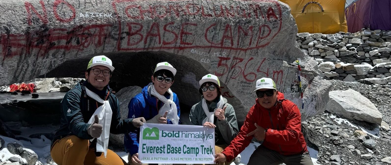 A group of trekkers posing with the Everest Base Camp sign in front of a boulder marked “Base Camp 5364m,” surrounded by snowy Himalayan peaks
