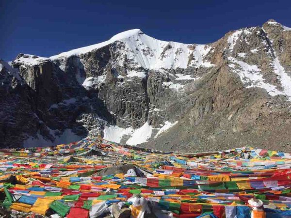 Colorful Tibetan prayer flags fluttering, with snow-covered Mount Kailash rising behind, the sacred Mount Kailash Mansarovar tour via Hilsa