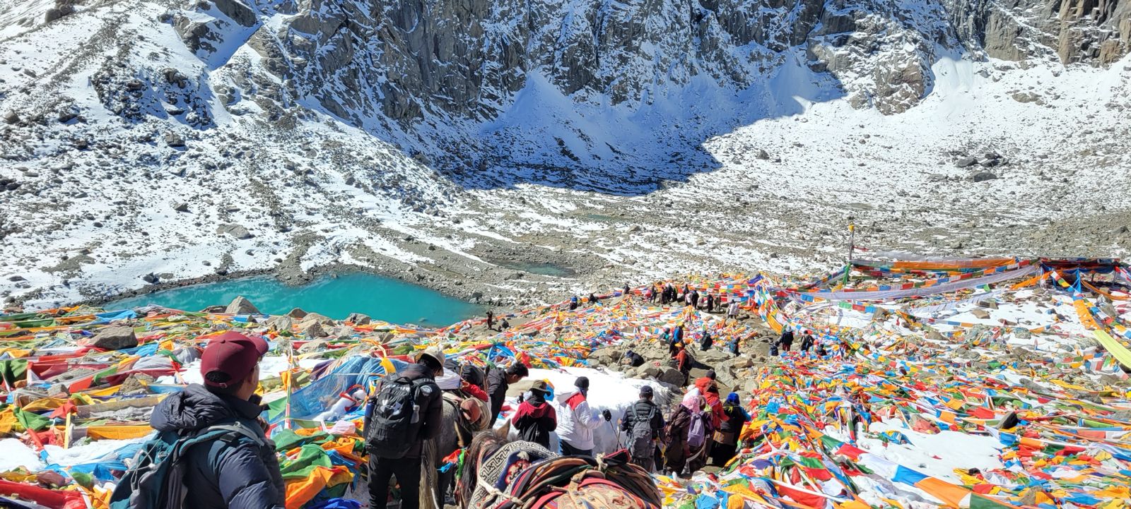 Pilgrims descending a snowy mountain slope during Kailash Yatra, surrounded by colorful prayer flags and a turquoise glacial lake
