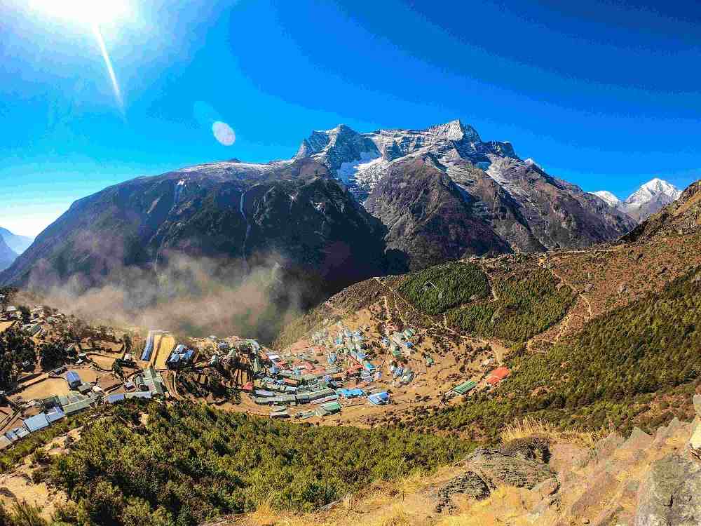 Aerial view of Namche Bazaar surrounded by towering Himalayan mountains under a bright blue sky during the Everest View Trek.