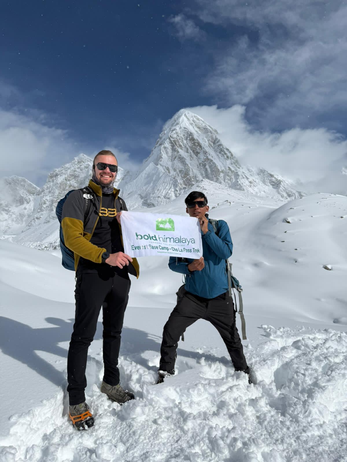 Two trekkers posing in deep snow during the Gokyo Lakes Renjo La Pass Trek in Nepal, holding a banner with a dramatic snow-covered Himalayan mountain rising behind them under a bright blue sky in the Everest region.