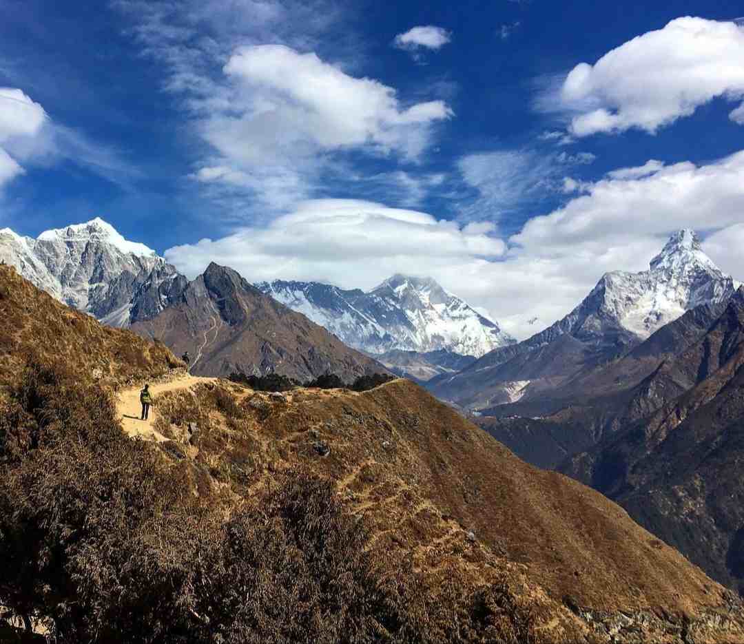 Trekker walking along a mountain trail with panoramic views of Mount Everest, Ama Dablam, and the Himalayas under a clear blue sky.