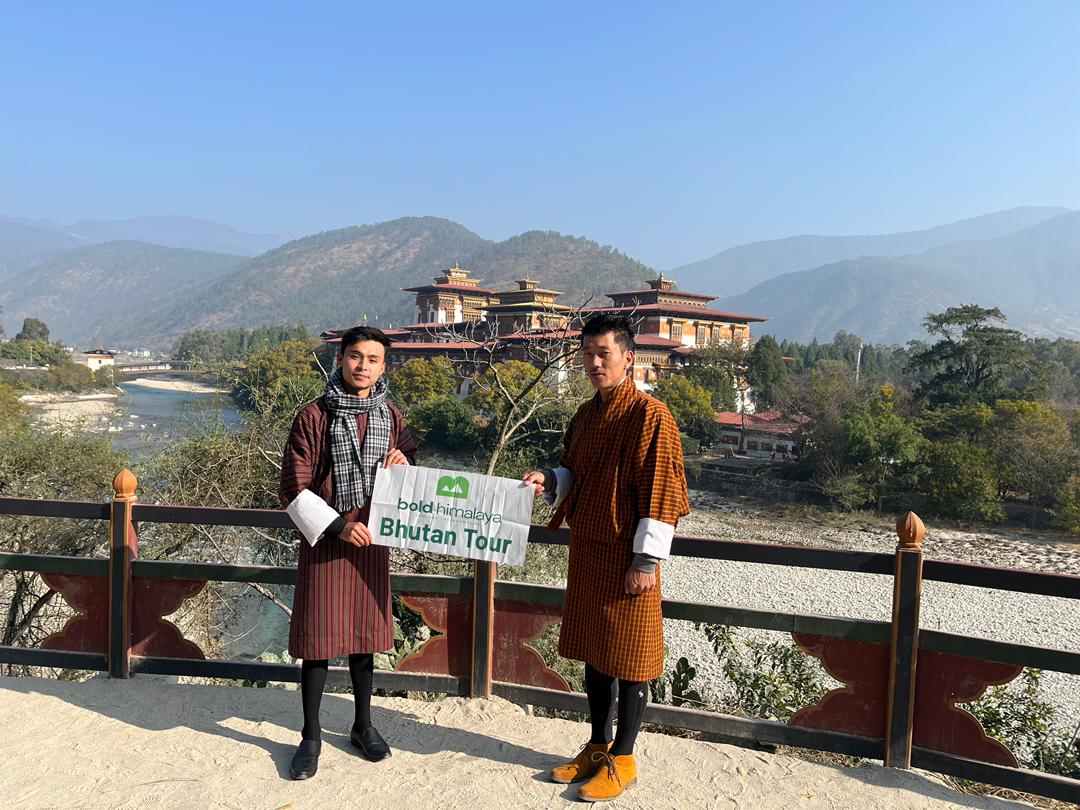 Two men in traditional Bhutanese dress holding a Bhutan Tour sign with Punakha Dzong and river valley in the background during a Bhutan Tour 5 Days 4 Nights.