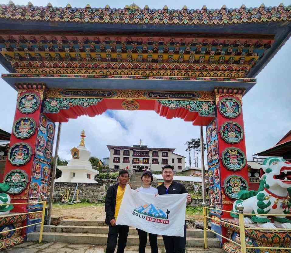 Trekkers holding a Bold Himalaya banner in front of the colorful Tengboche Monastery gate with Buddhist art and mountain backdrop during the Everest View Trek.