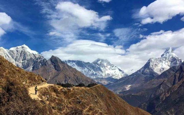 Trekker walking on the Everest Base Camp trail with snow-capped Himalayan mountains, including Mount Everest, under a bright blue sky.
