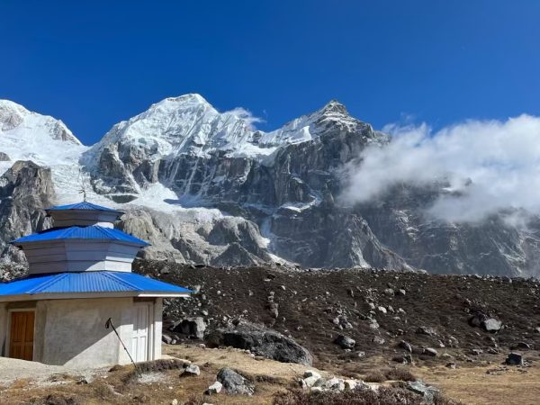 A small white temple structure with a blue roof stands beneath towering snow-covered peaks and rocky slopes along the Kanchenjunga Base Camp Trek