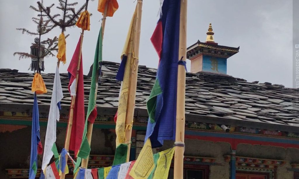 Colorful prayer flags fluttering beside a traditional monastery building along the Tsum Valley Trek, with a stone-roofed architecture in the background