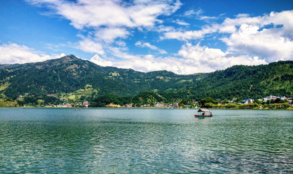Serene view of Phewa Lake in Pokhara with calm water, green hills, scattered boats, and blue sky with clouds, one of the best tourist places to visit in Nepal