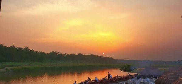 Golden sunset over a calm river in Nepal, with people relaxing along the riverbank, reflecting a peaceful and luxurious riverside experience