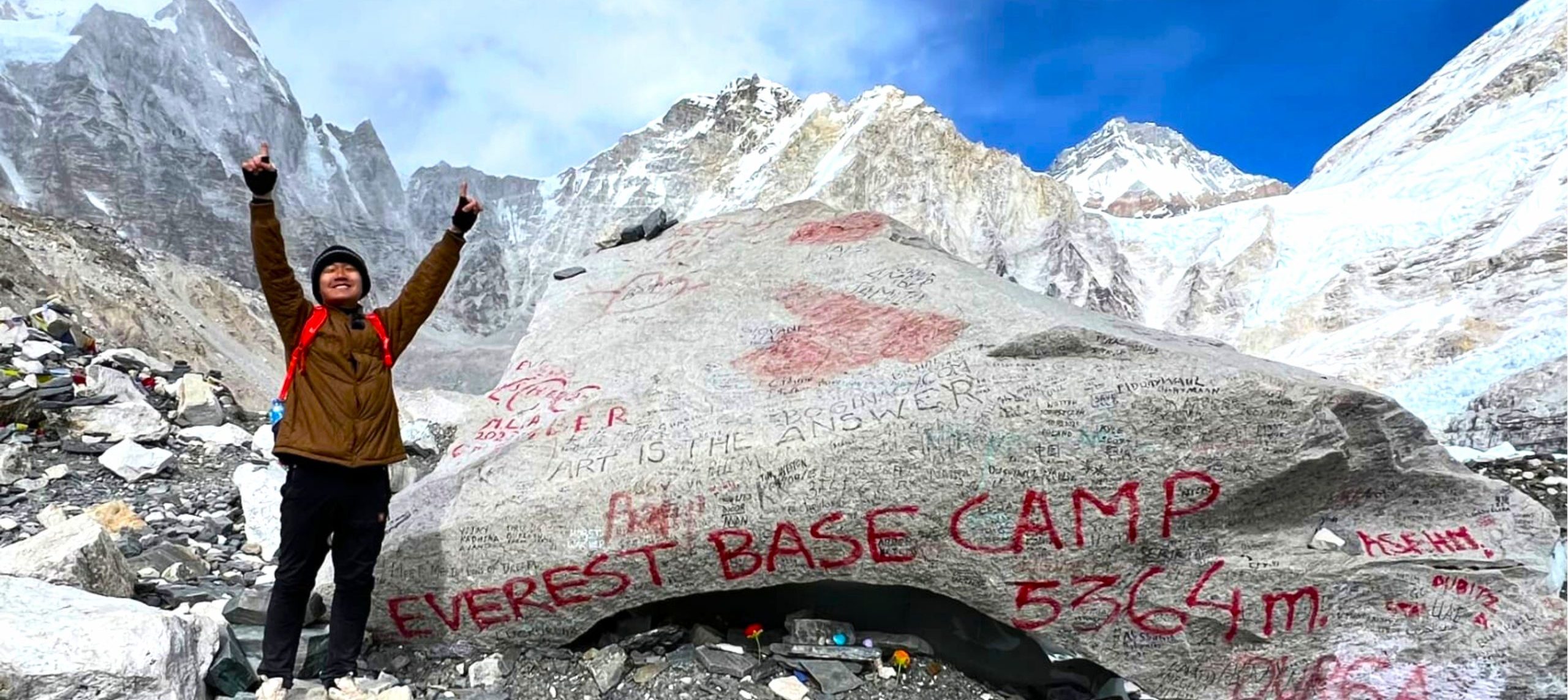 Trekker celebrating at Everest Base Camp beside the iconic rock marker with snow-covered Himalayan peaks in the background, the best season for the Everest Base Camp Trek