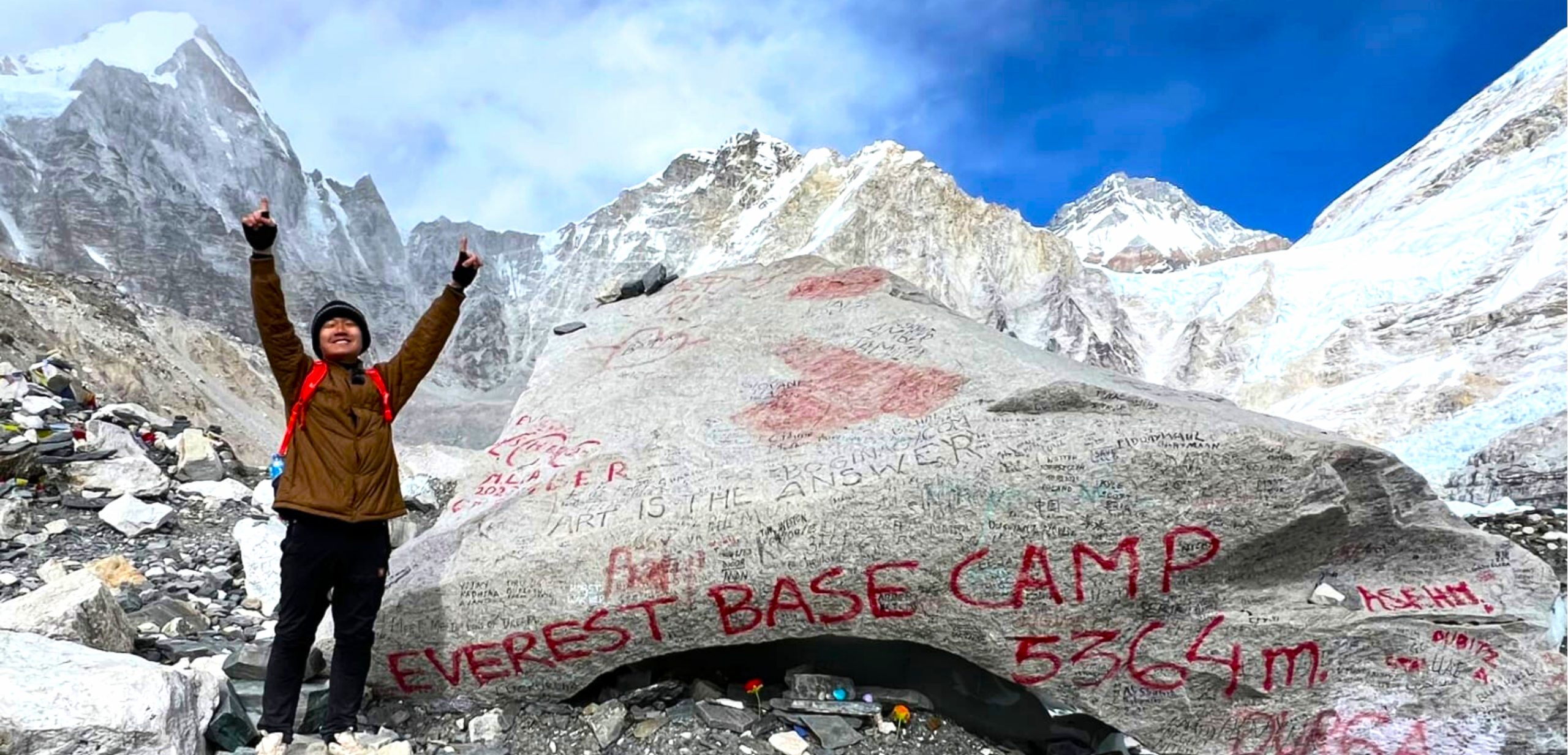 Trekker celebrating at Everest Base Camp beside the iconic rock marker with snow-covered Himalayan peaks in the background, the best season for the Everest Base Camp Trek