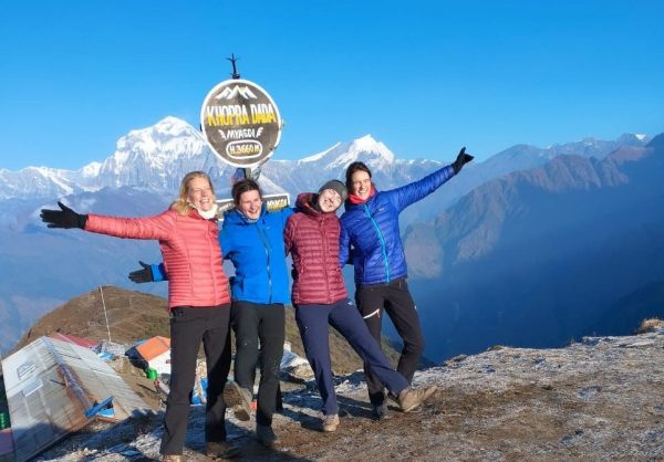Four trekkers in colorful down jackets joyfully pose at the Khopra Danda viewpoint with snow-capped Himalayan peaks glowing under the clear blue sky-best treks in nepal