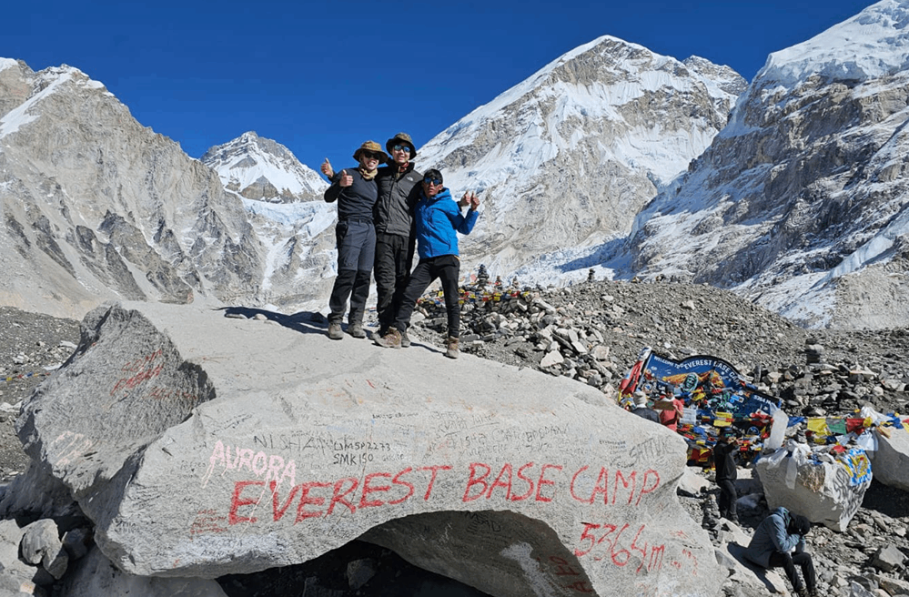 Bold Himalaya trekking team posing with the Everest Base Camp sign, surrounded by snowy peaks and prayer flags.