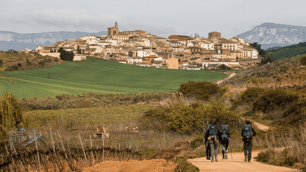 Hikers walking along the Camino de Santiago trail toward a traditional Spanish village surrounded by rolling countryside