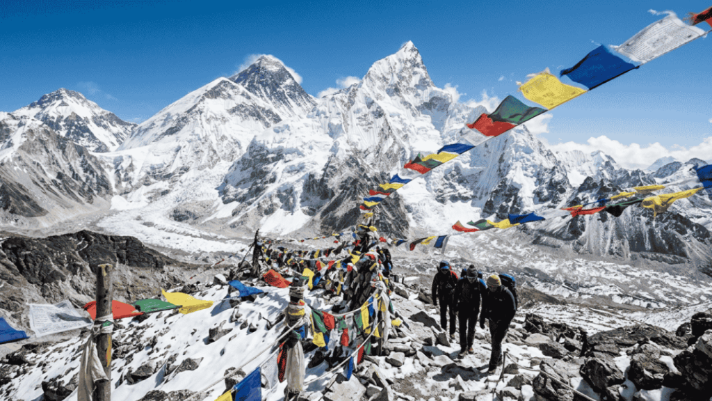 Trekkers on a rocky Himalayan path lined with prayer flags, facing Mt. Everest.