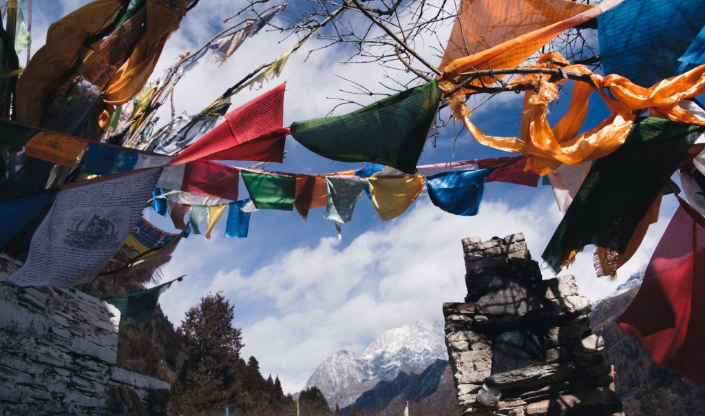 Prayer flags flutter across a mountain pass with snow-capped Himalayan peaks in the background.